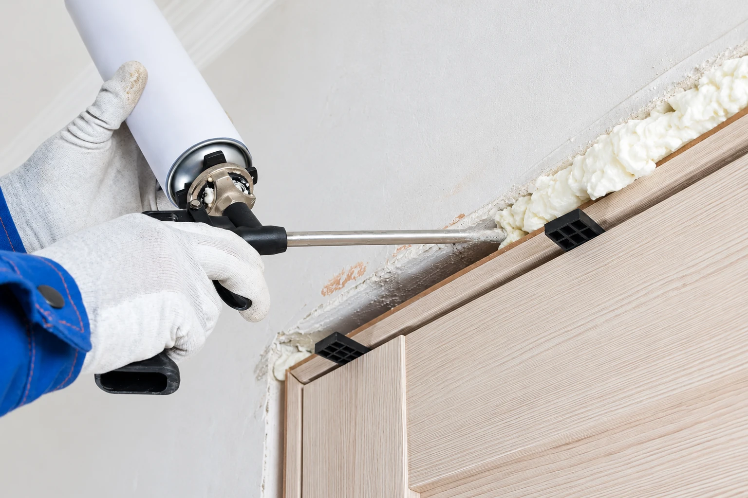 Gloved worker applies expanding foam around the edge of a wall opening with a spray gun.