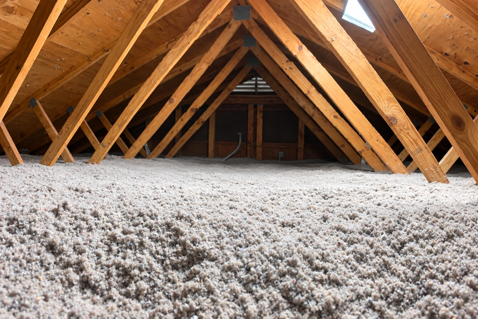 Loose-fill attic insulation covers the floor beneath a framework of exposed wooden roof trusses.