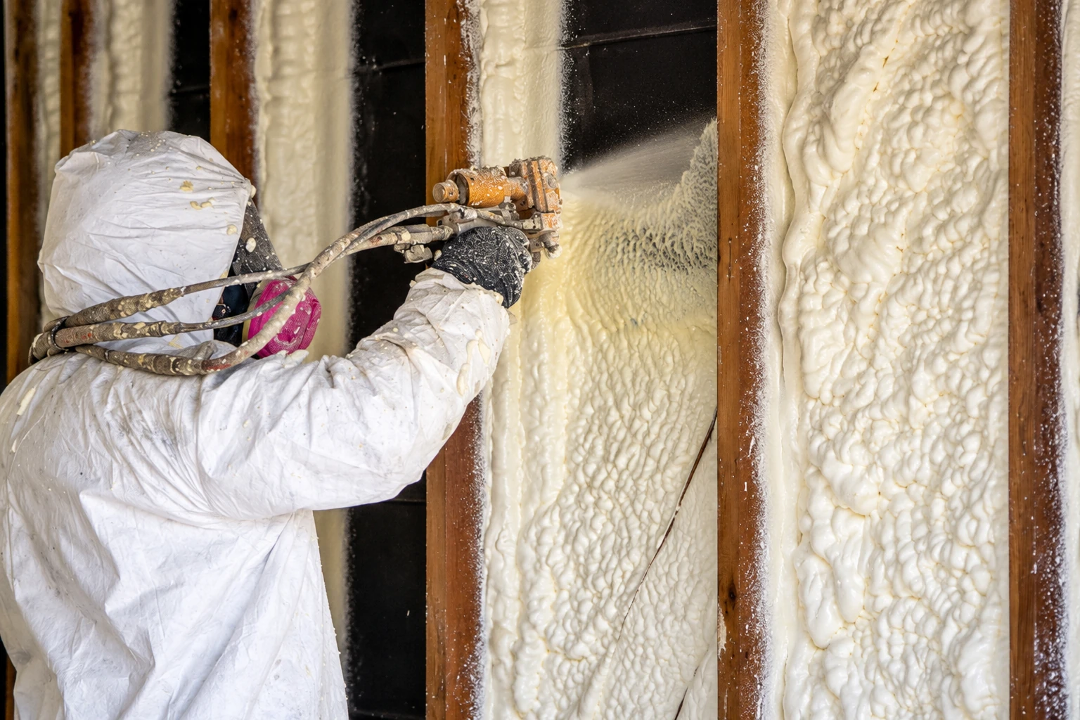 Worker in protective clothing sprays foam insulation between wooden wall studs.