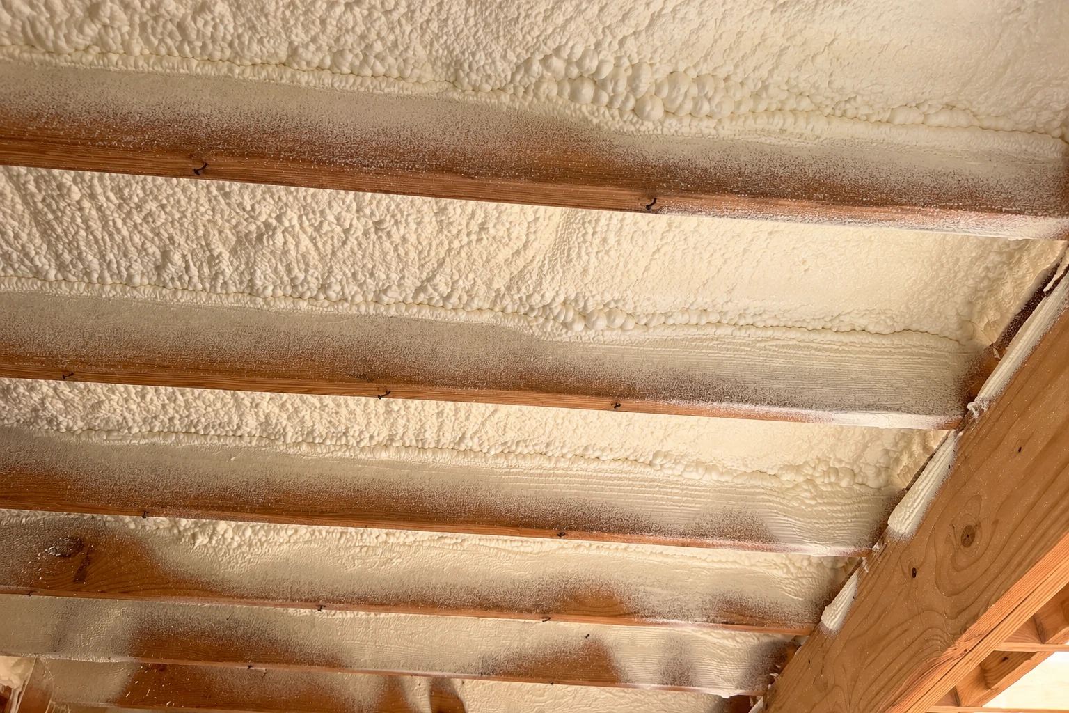 White spray foam insulation fills the spaces between wooden rafters on the underside of a roof.