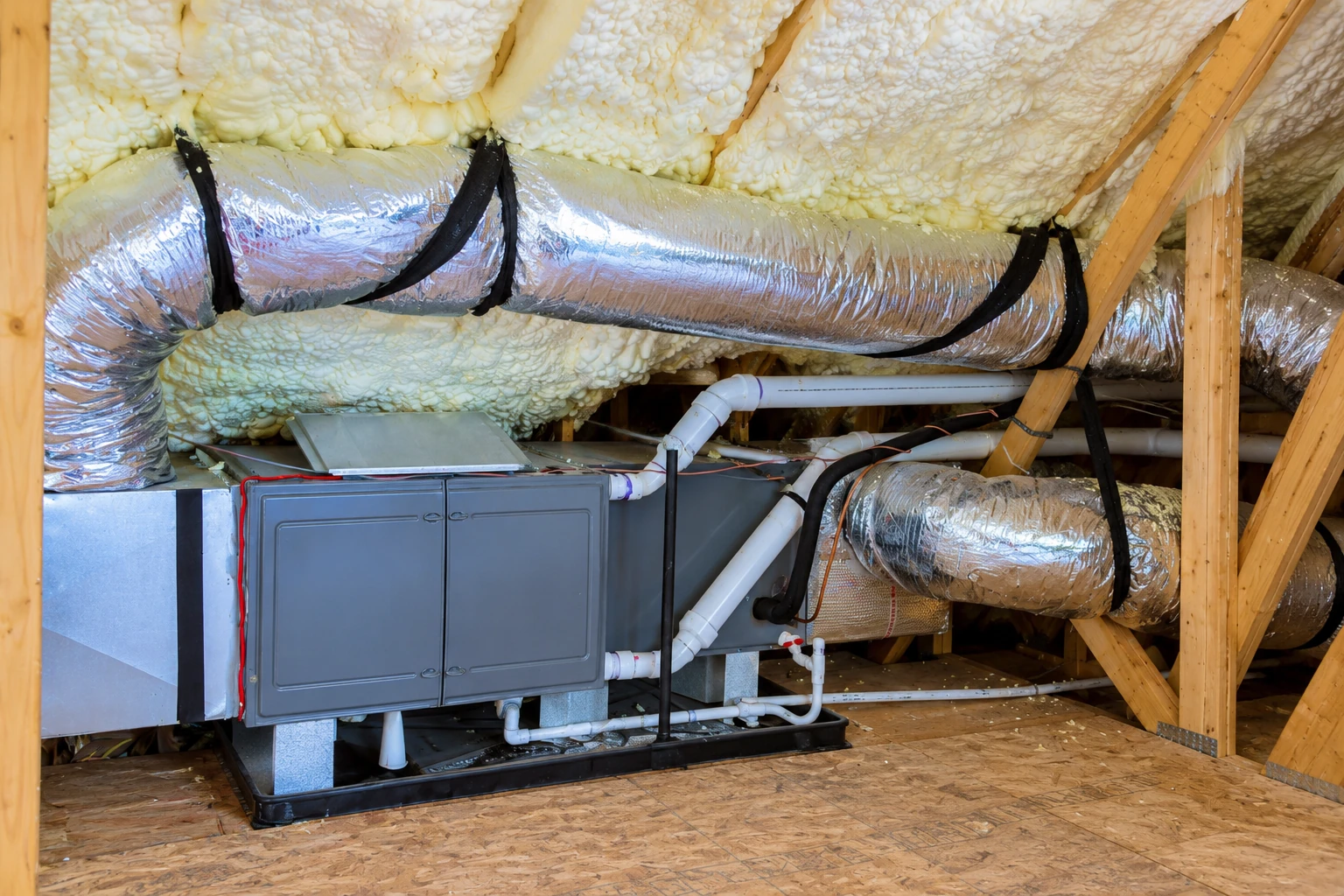 Attic HVAC unit and silver ducts beneath thick cream-colored spray foam insulation.
