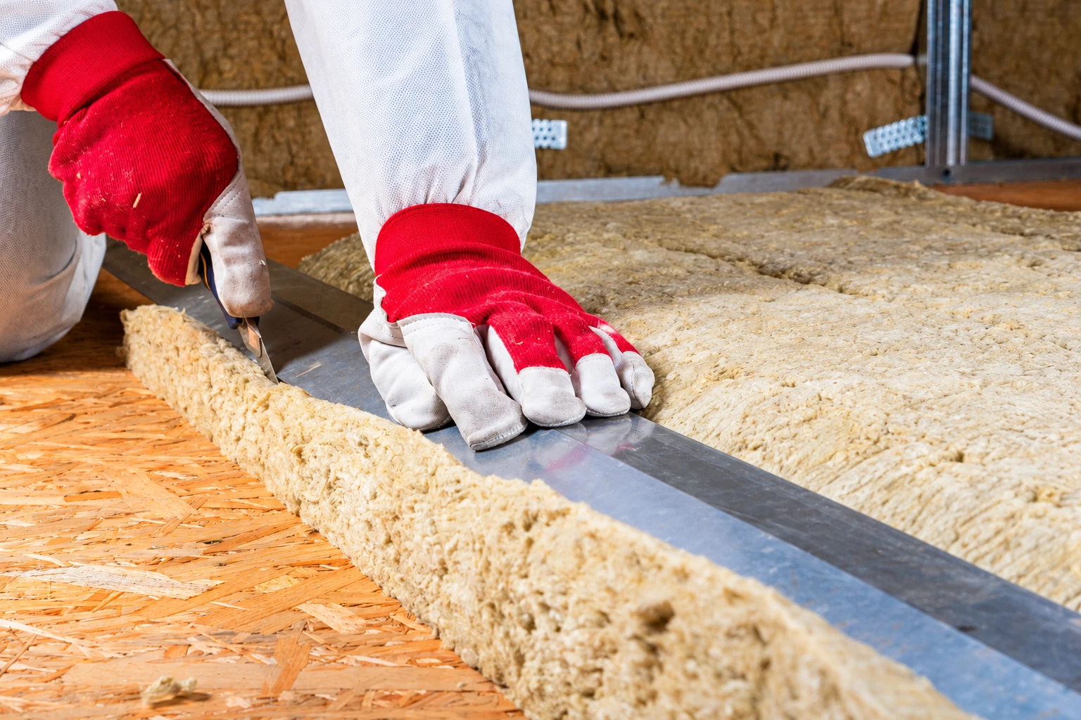 A worker in red-and-white gloves cuts a batt of yellow insulation beside metal framing on a plywood floor.