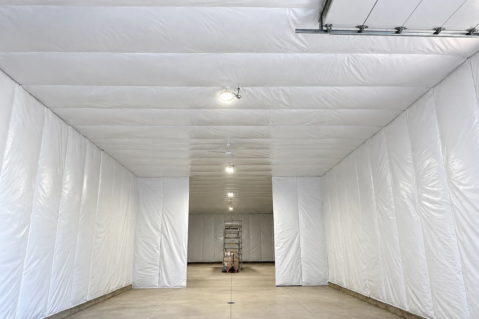 A garage interior is lined with white vapor-barrier sheeting across its walls and ceiling, with a ladder at the far end.