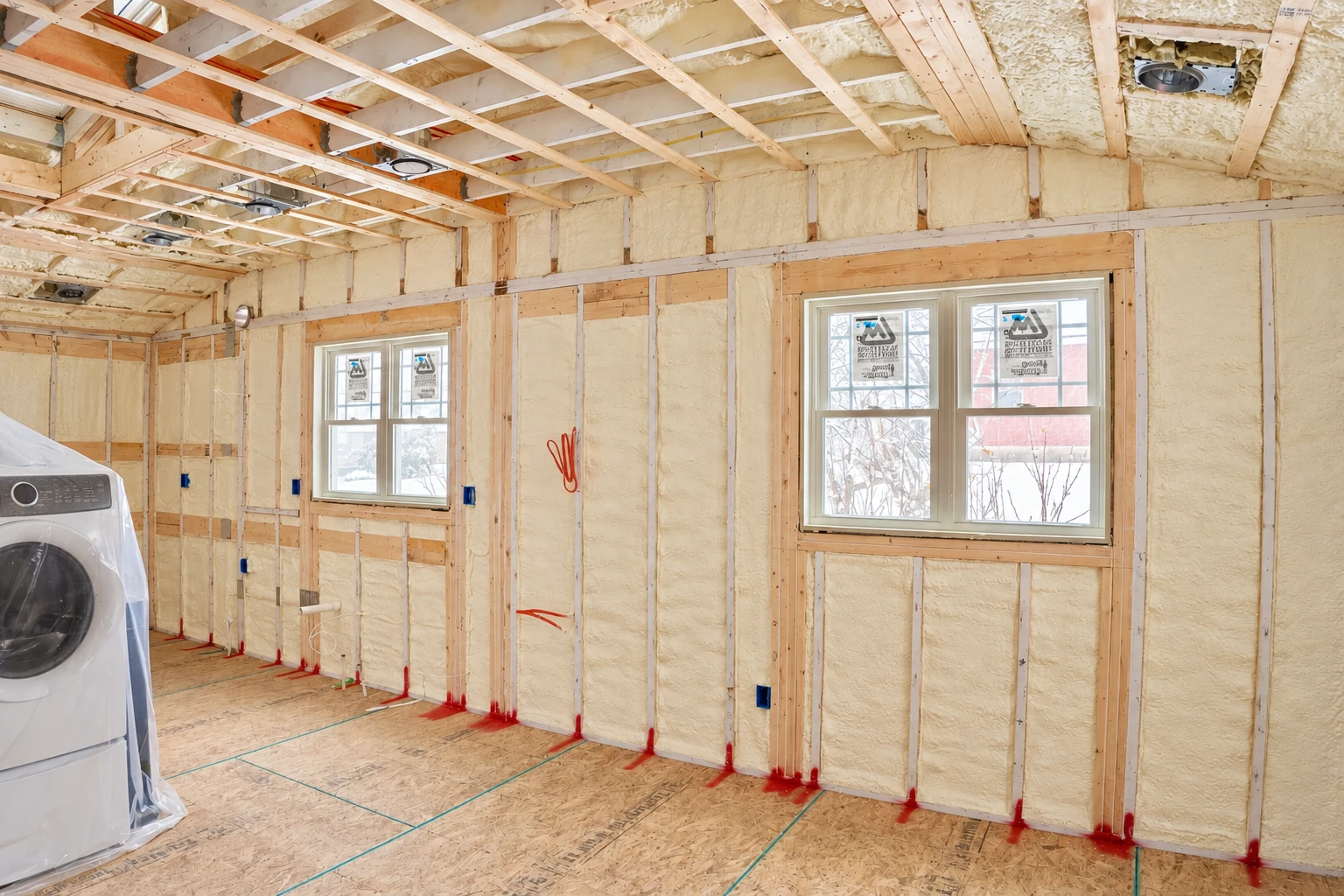 Unfinished room with pale spray foam insulation between wall studs, two windows, and exposed ceiling framing.